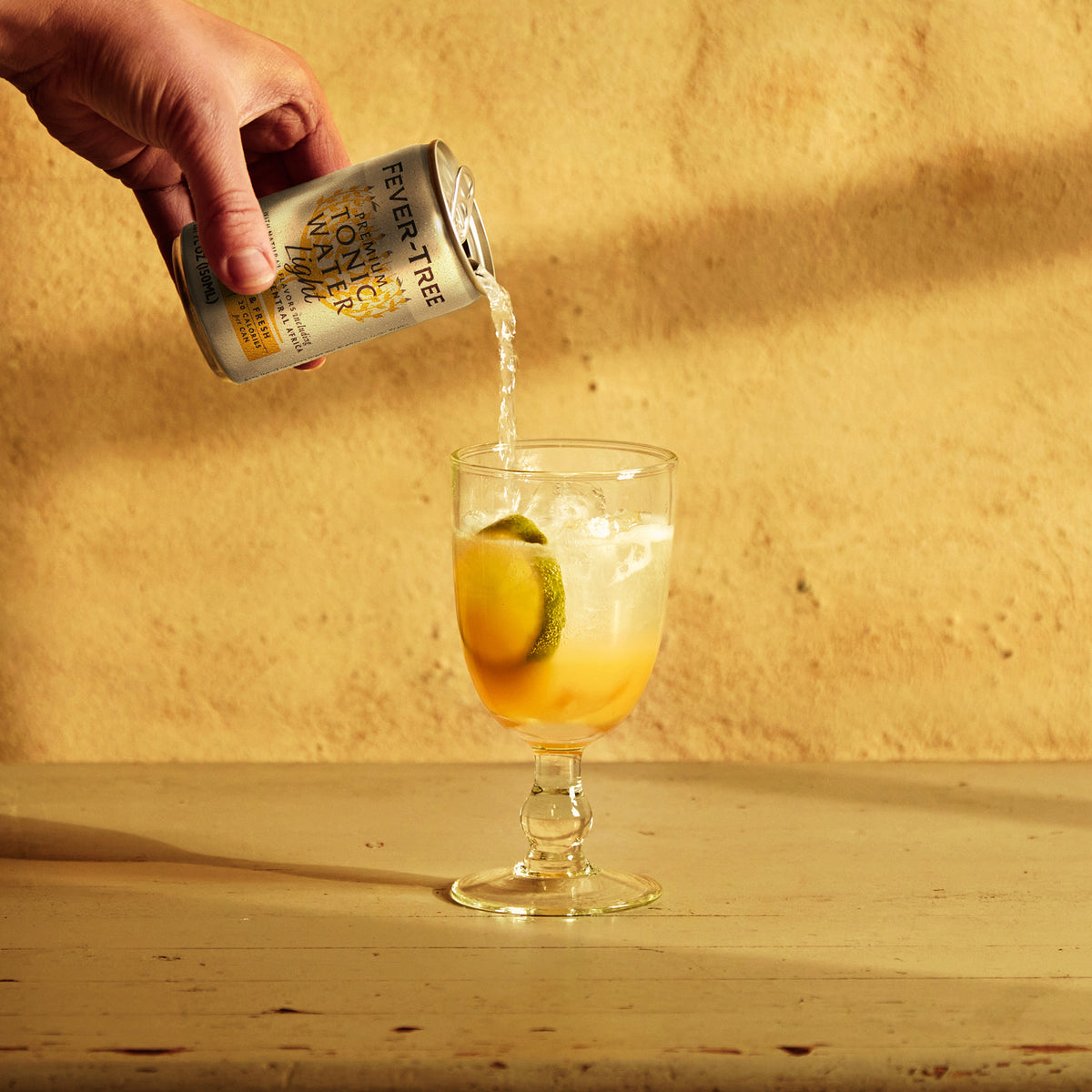 A hand pours Mother Root Fever Tree Light Premium Tonic [8 x 5.07 fl oz] over ice, lemon, and lime in a glass.