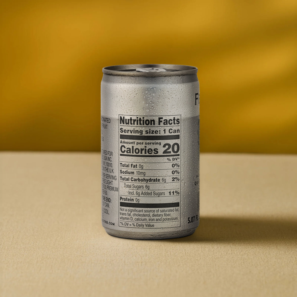 A silver can of Mother Root Fever Tree Light Premium Tonic [8 x 5.07 fl oz] with condensation rests on a beige surface before a yellow background, displaying a nutrition label with 20 calories per can.