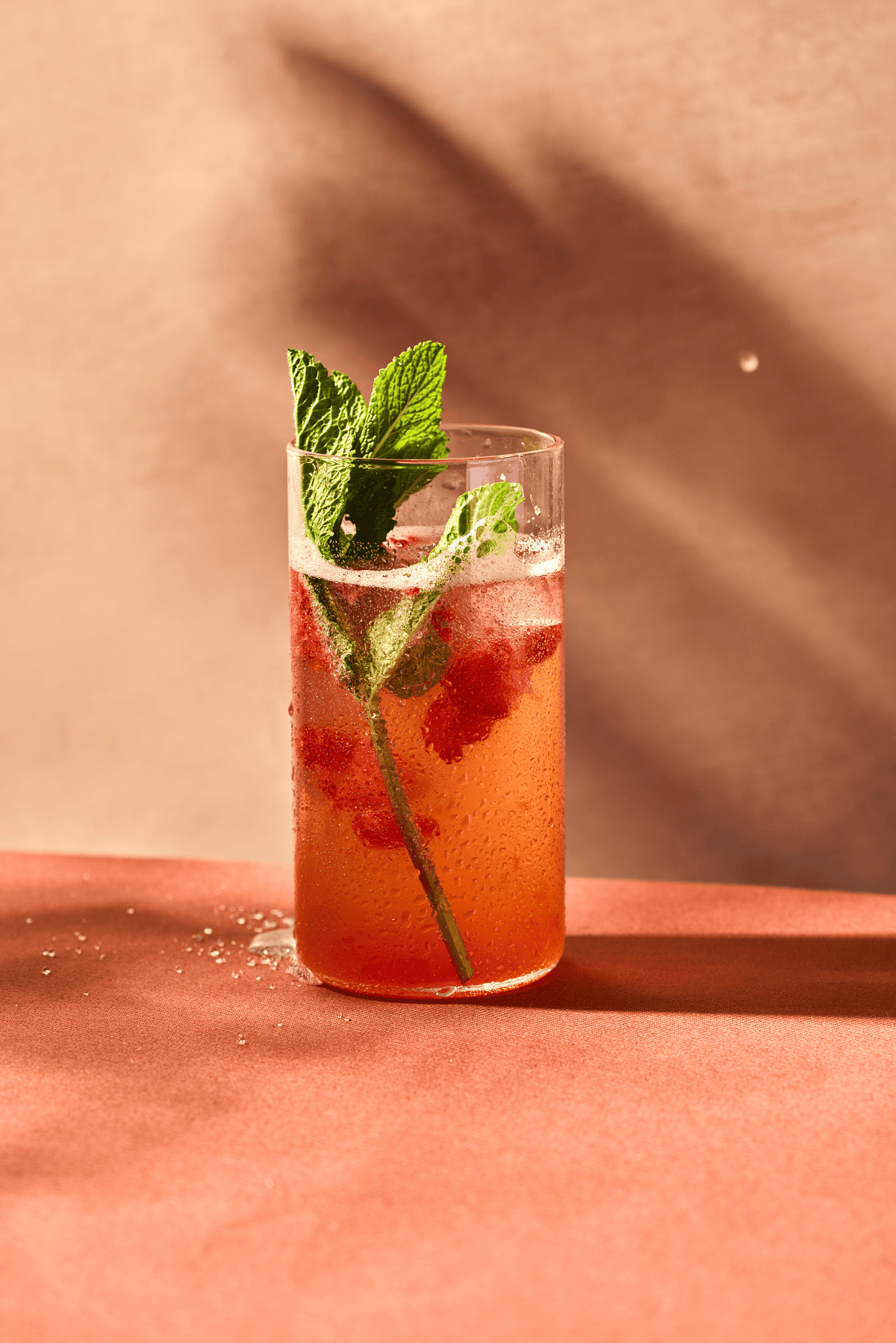 A refreshing red drink in a clear glass, garnished with mint leaves, set against a soft pink background with gentle shadows, exuding a summery vibe.