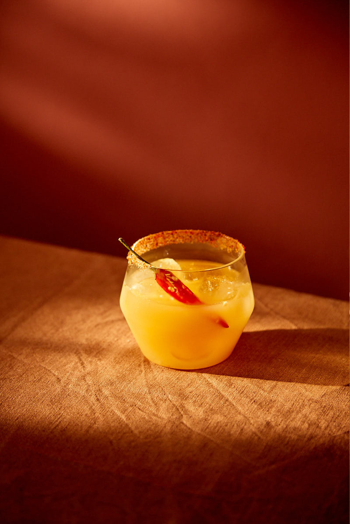 A Mother Root cocktail in a glass with a spicy rim sits on a textured surface. The drink is orange-yellow, garnished with a red chili pepper, under warm ambient lighting.