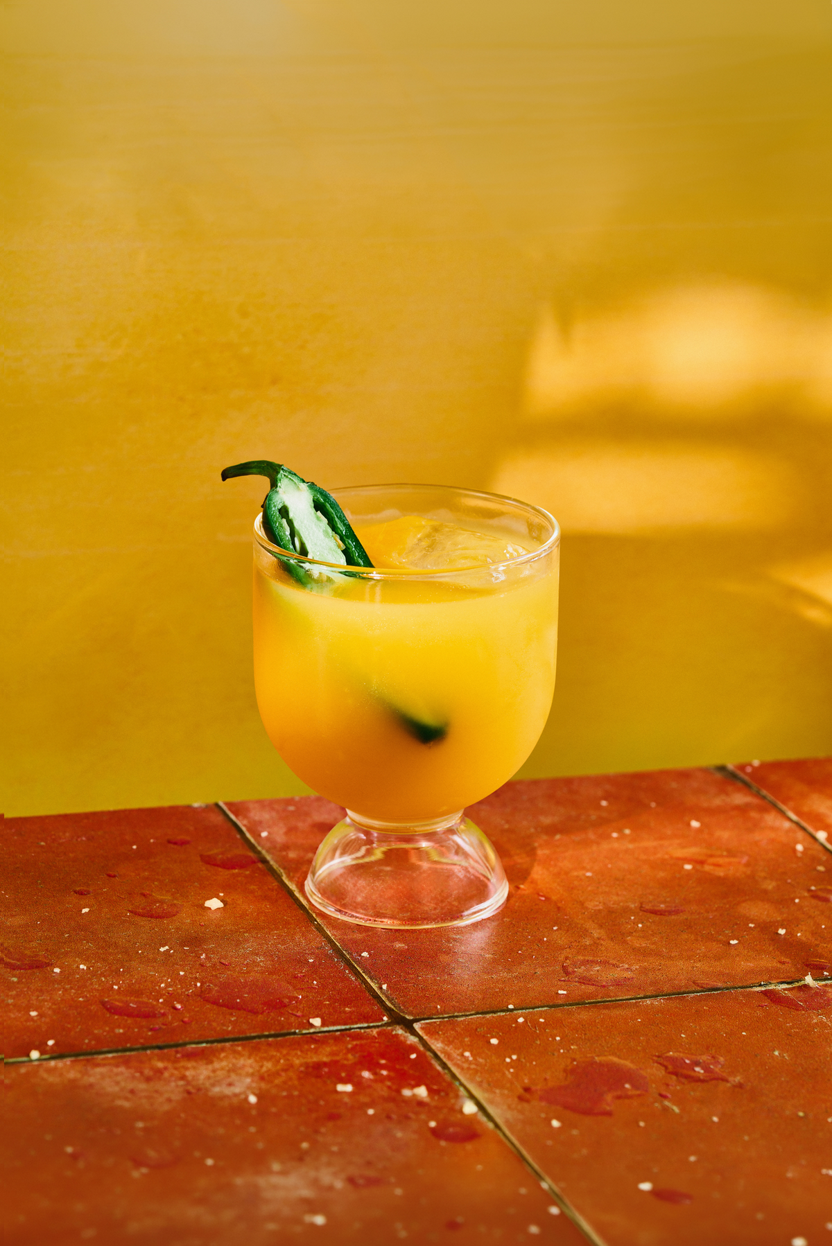 Golden cocktail in a pedestal glass with ice and fresh jalapeno, set against a vibrant yellow background and terracotta tile surface.