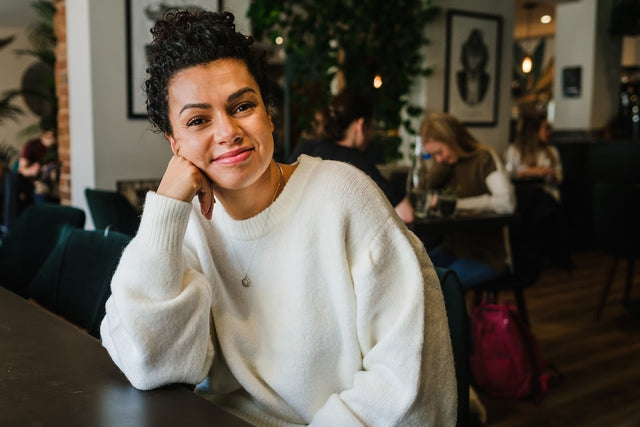 Woman with curly hair in a white sweater smiling and sitting at a table in a cozy latte café.