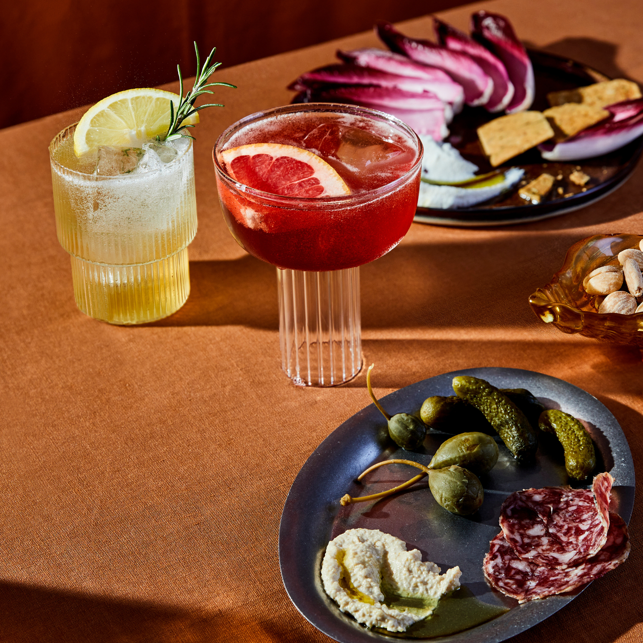 Sip two cocktails and appetizers with Mother Root’s Limited Edition Triple Bottle Set on an orange tablecloth.