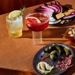 Sip two cocktails and appetizers with Mother Root’s Limited Edition Triple Bottle Set on an orange tablecloth.