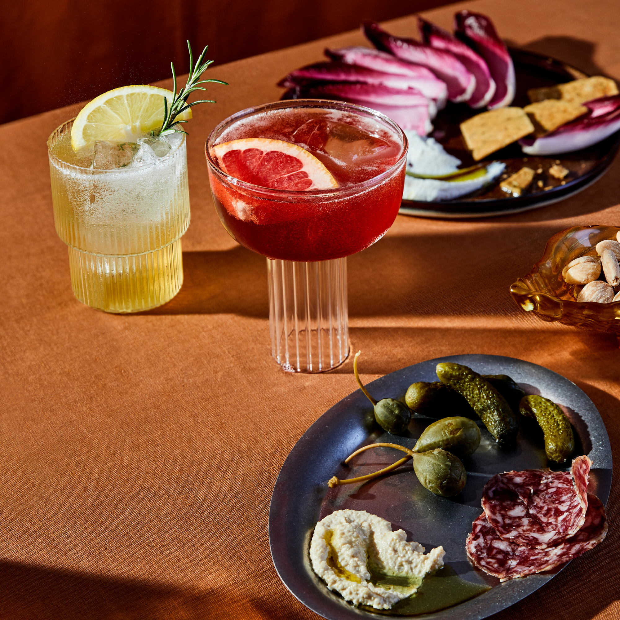 Sip two cocktails and appetizers with Mother Root’s Limited Edition Triple Bottle Set on an orange tablecloth.