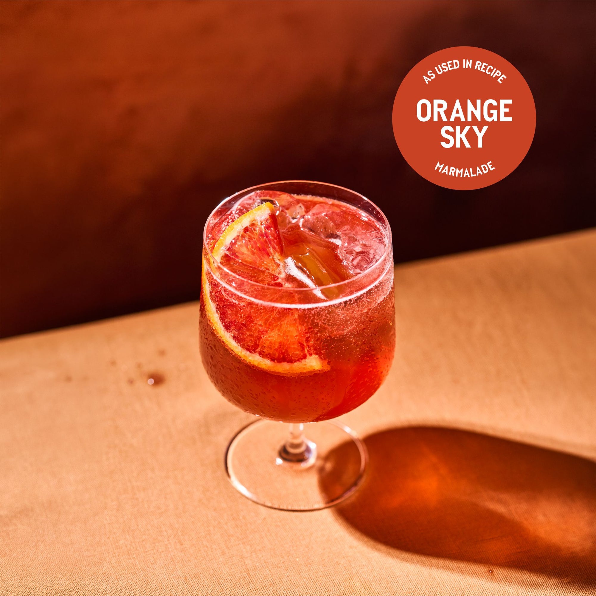 A deep red-orange cocktail served over ice in a stemmed glass, garnished with a blood orange slice, on a warm terracotta surface in golden light. A circular badge reads 'As used in recipe: Orange Sky Marmalade