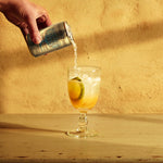 A hand pours Mother Root Refreshingly Light Tonic Water into a glass with ice, lemon, and lime on a wooden table.