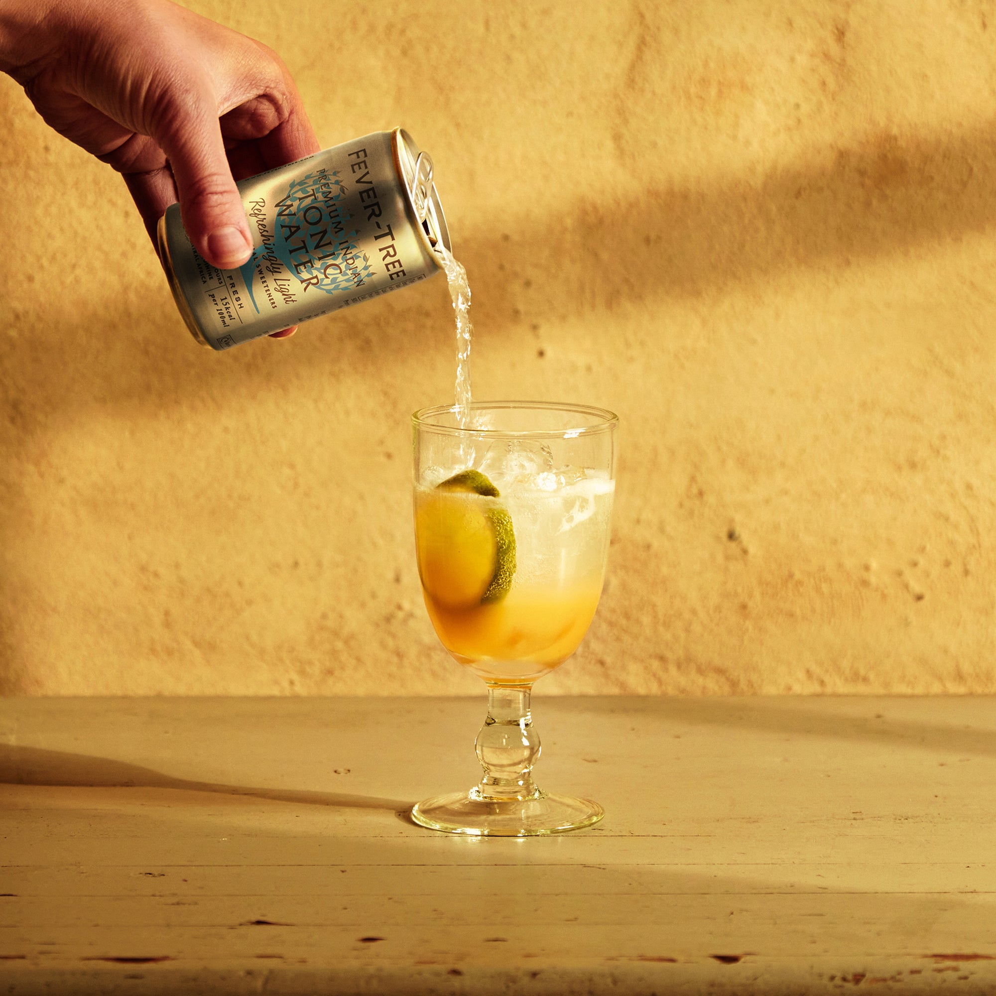 A hand pours Mother Root Refreshingly Light Tonic Water into a glass with ice, lemon, and lime on a wooden table.
