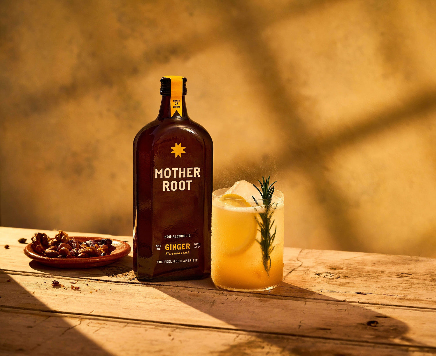 A bottle of Mother Root ginger, ice with rosemary, and a small plate of nuts on a rustic wooden table.