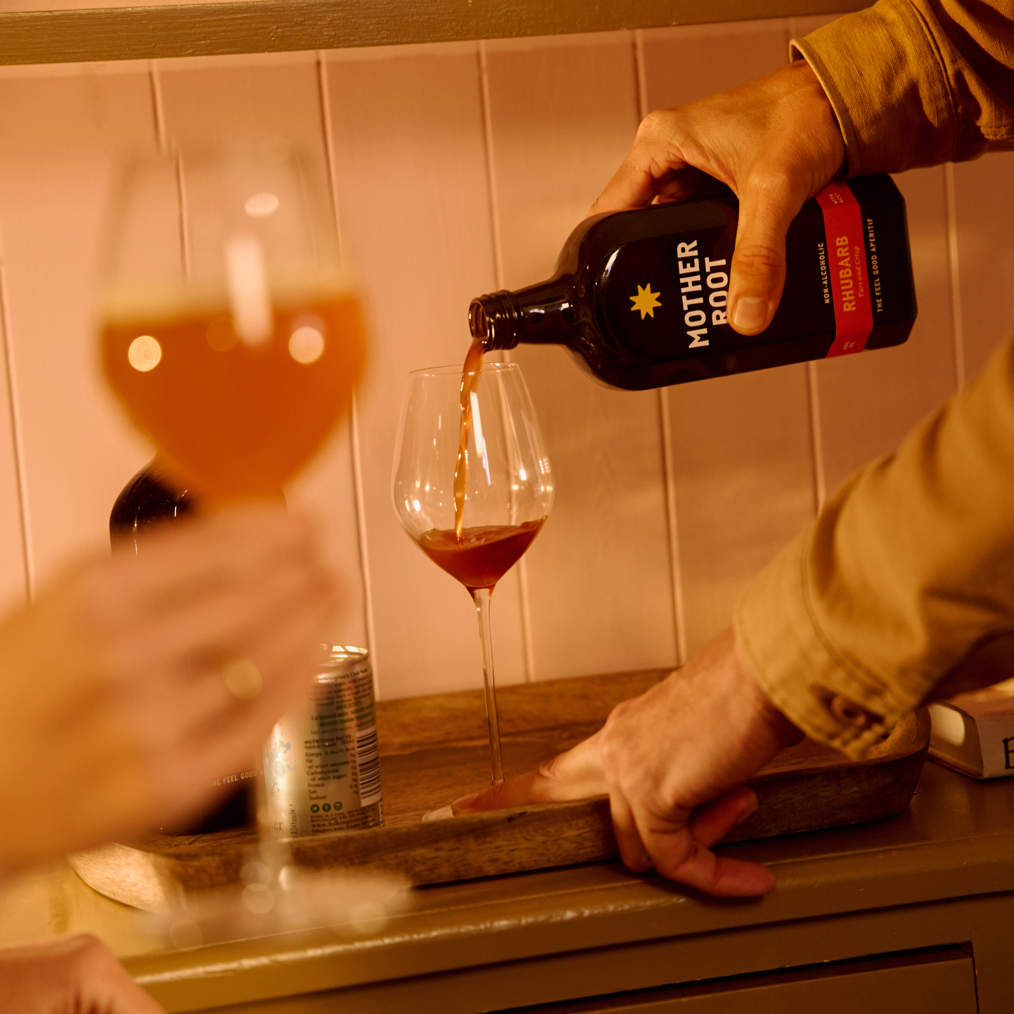A hand pours a dark liquid from a "Mother Root" bottle into a wine glass on a tray, with another blurred glass in the foreground, creating a cozy, warm ambiance.