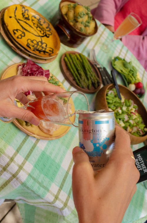 A hand pours sparkling water from a can into a glass on a table with fresh salads and roasted potatoes.