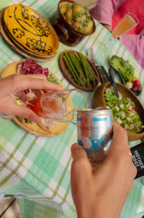 A hand pours sparkling water from a can into a glass on a table with fresh salads and roasted potatoes.