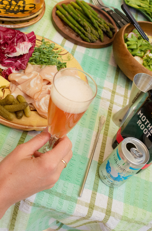 A hand holds a foamy drink above a table with vegetables, charcuterie, and beverages on a green checked cloth.
