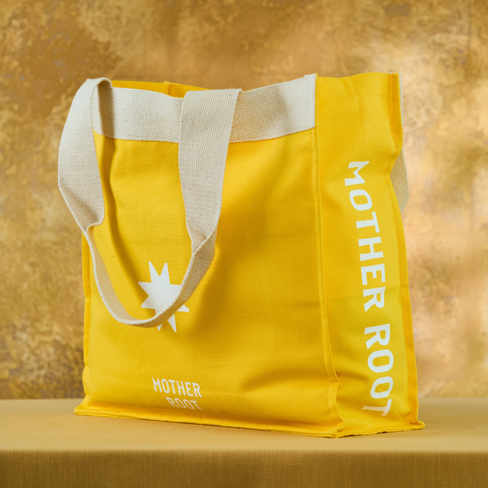 A bright yellow reusable tote bag with beige canvas handles, featuring white printed text reading 'MOTHER ROOT' vertically on the side and a white starburst logo near the bottom, set against a warm textured gold background.