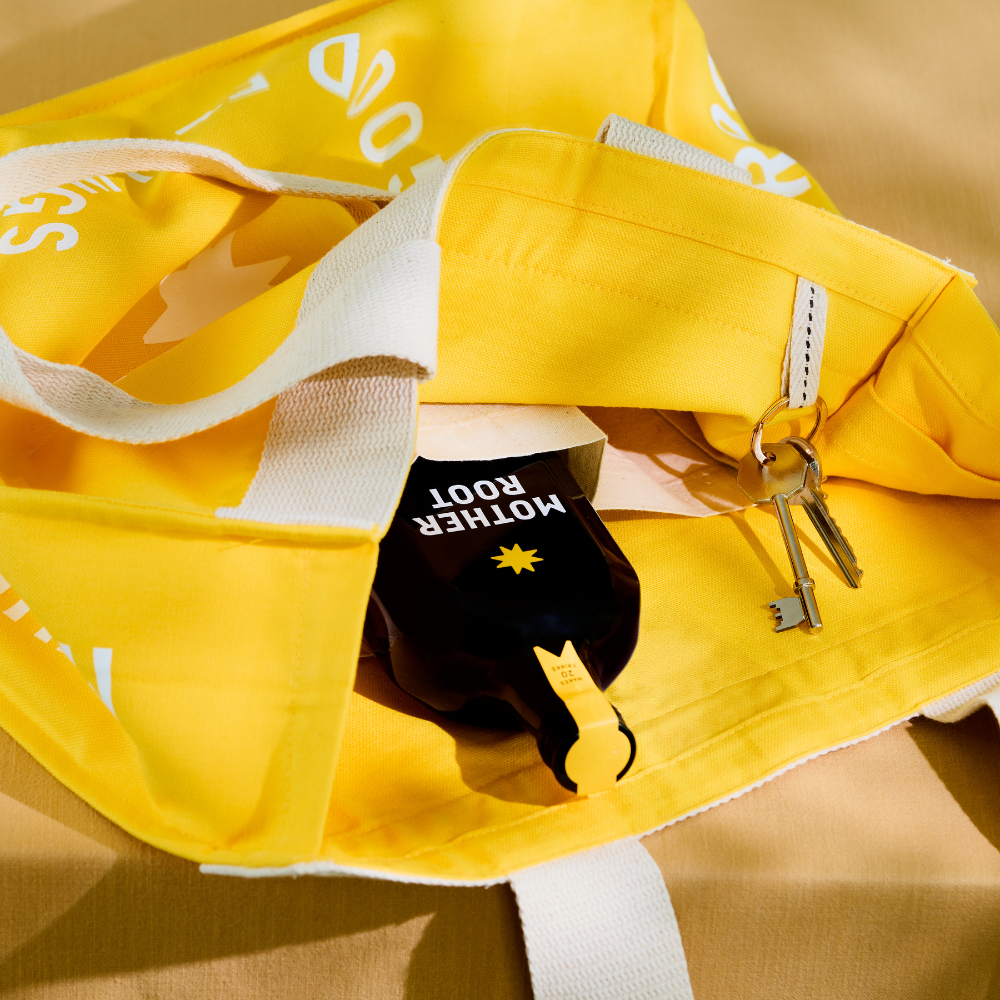 A yellow Tote Bag by Mother Root with side pockets holds a bottle and silver keys on a tan surface.