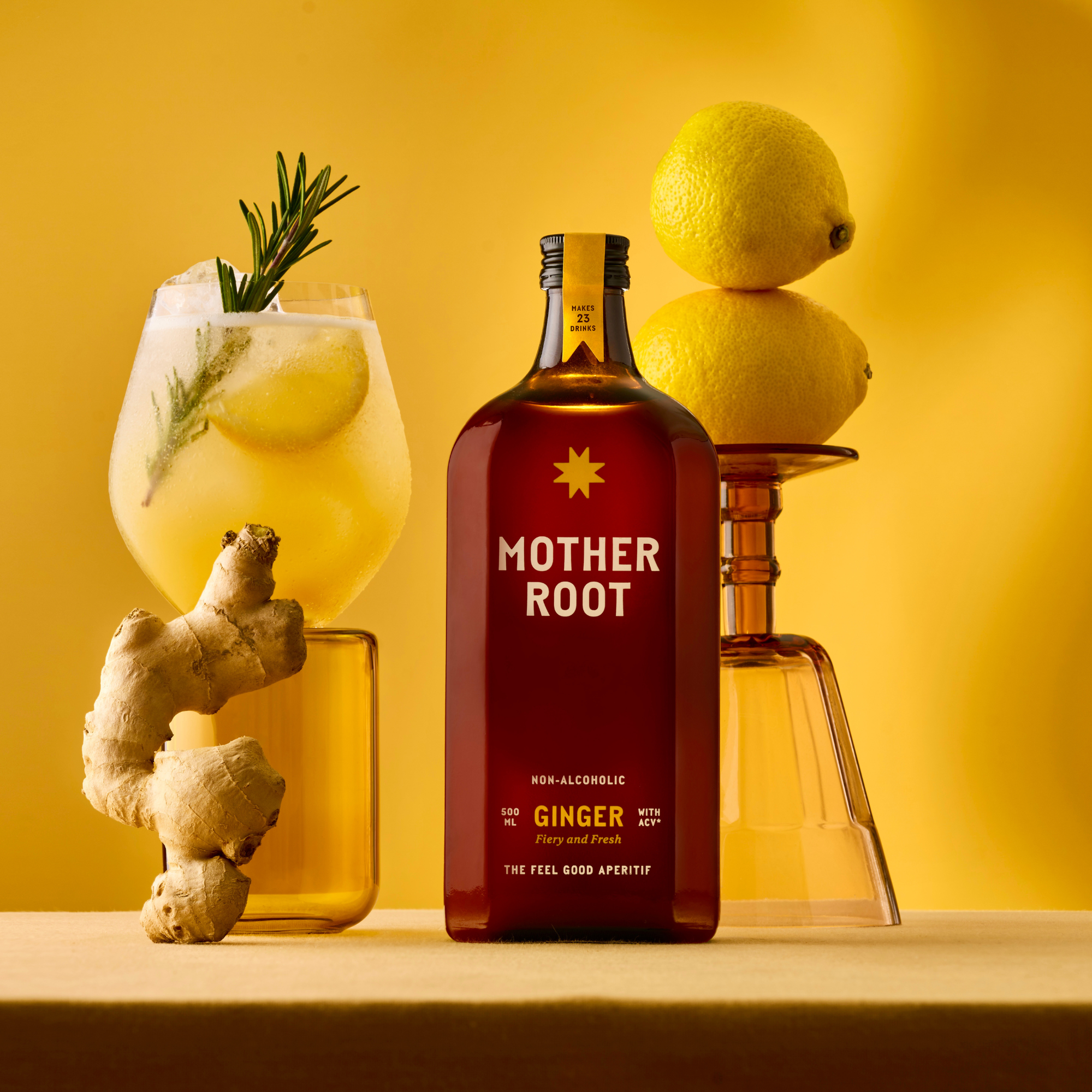 Bottle of 'Mother Root' ginger drink with a serve with rosemary, lemons and ginger on a yellow background.