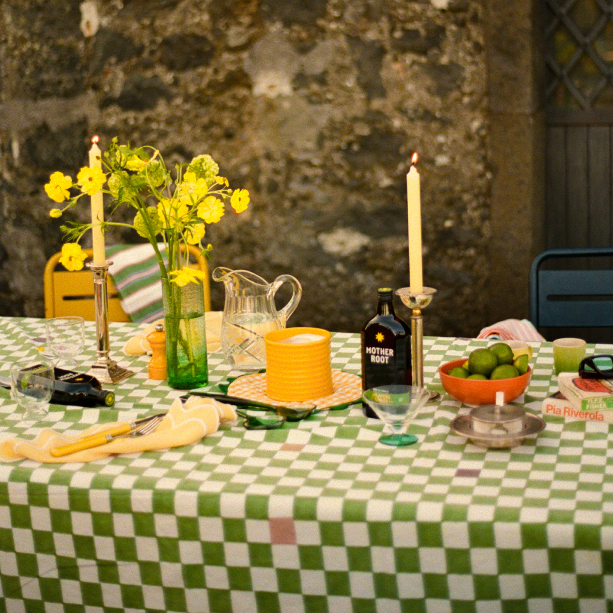 A checkered tablecloth with a cozy, rustic vibe features bright yellow flowers, lit candles, citrus in a bowl, and a bottle labeled "Mother Root."