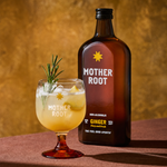 A brown bottle labeled "Mother Root" with a yellow star stands next to a glass of ginger drink, garnished with rosemary and ice, on a warm, golden background.