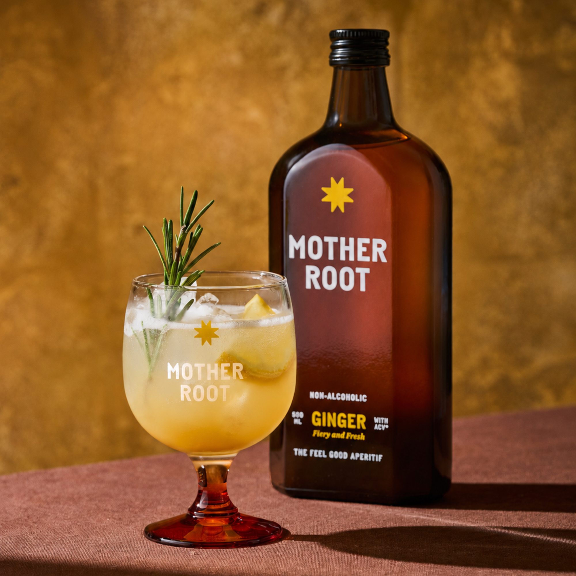 A brown bottle labeled "Mother Root" with a yellow star stands next to a glass of ginger drink, garnished with rosemary and ice, on a warm, golden background.