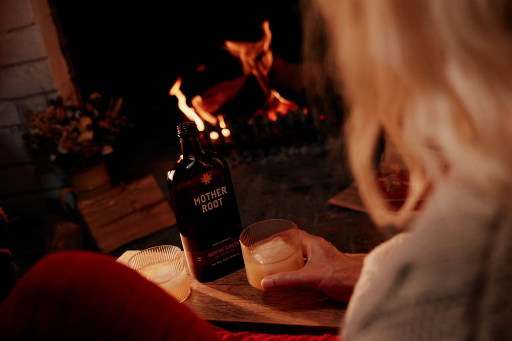A hand reaches for a glass of Bonfire Ginger beside the bottle, fire burning behind