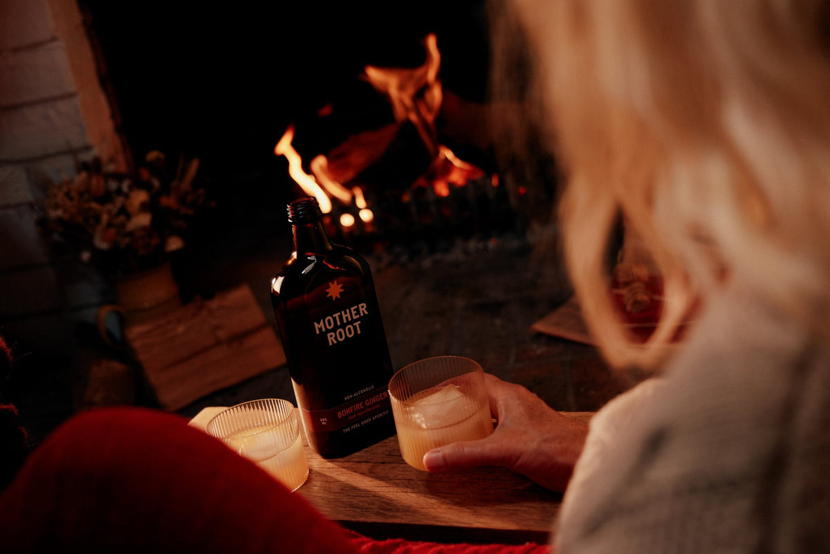 A hand reaches for a glass of Bonfire Ginger beside the bottle, fire burning behind