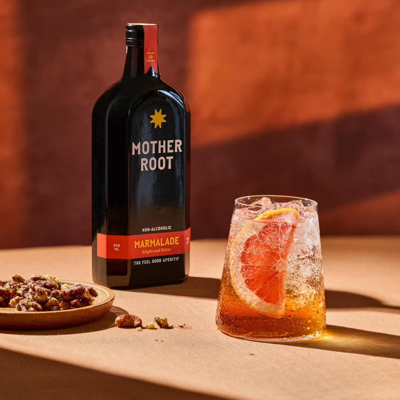 Mother Root Marmalade 500ml bottle beside a tall glass of Mother Root spritz garnished with grapefruit, with a plate of candied nuts, lit by warm low sun against a terracotta backdrop