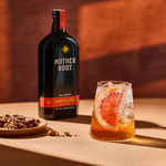 Mother Root Marmalade 500ml bottle beside a tall glass of Mother Root spritz garnished with grapefruit, with a plate of candied nuts, lit by warm low sun against a terracotta backdrop