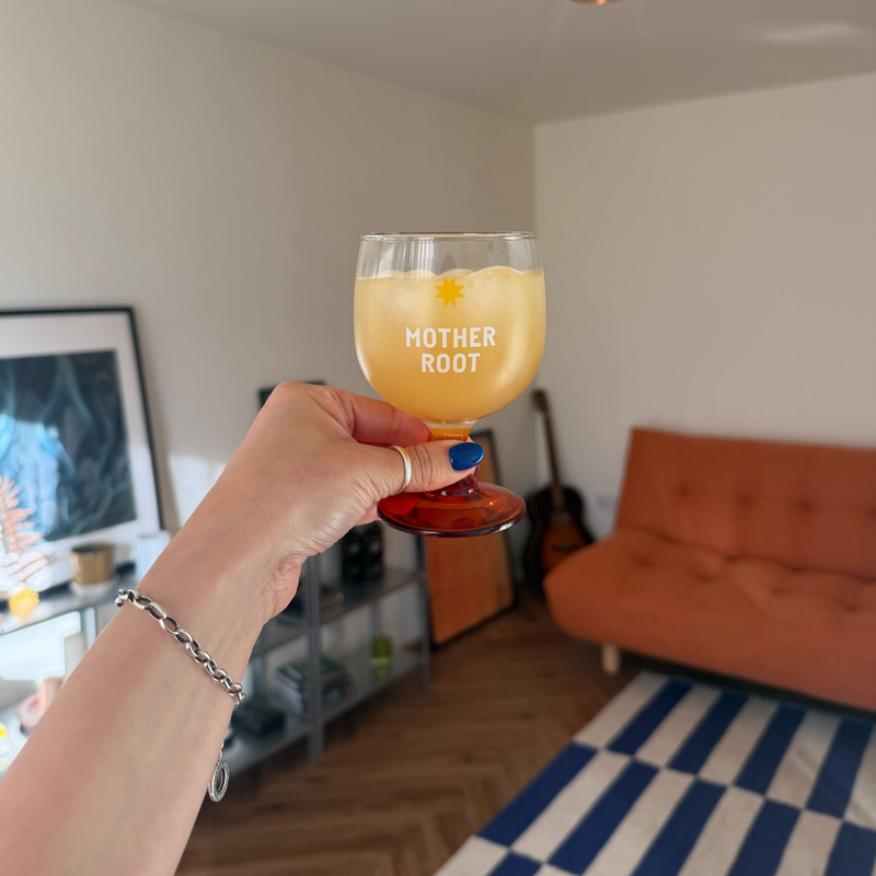 A hand with blue nails holds a "Mother Root" glass of orange drink in a cozy, modern living room. A striped rug, sofa, and guitar are visible.