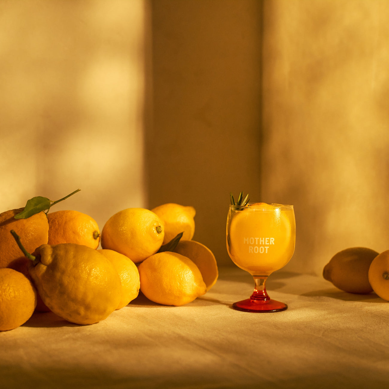A group of lemons is arranged on a sunlit table beside a glass of yellow liquid with "Mother Root" written on it, creating a warm, fresh ambiance.