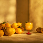A group of lemons is arranged on a sunlit table beside a glass of yellow liquid with "Mother Root" written on it, creating a warm, fresh ambiance.