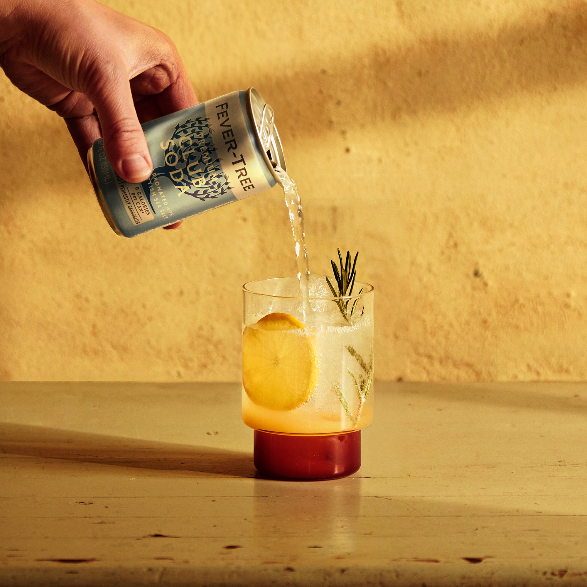A hand pours Fever-Tree tonic water into a glass with a large lemon slice and rosemary, on a wooden table against a warm, textured wall backdrop.