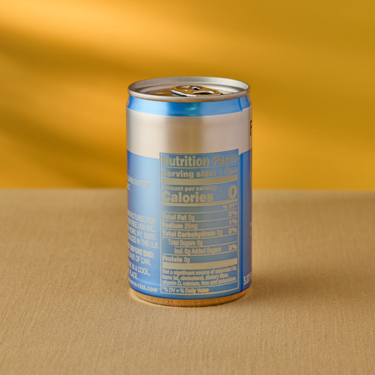 A small blue and silver can with nutrition facts is placed on a beige surface against a mustard yellow background. The can emphasizes zero calories.