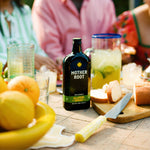 Bottle of Mother Root Jalapeño Ginger sits on a table amid a vibrant outdoor gathering, surrounded by citrus fruits, bread, and lemonade. Warm, inviting atmosphere.