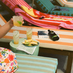 Colorful poolside scene with striped lounge chairs and towels. A hand holds a cocktail, surrounded by drinks, a plate of fruit, and books. Relaxing, vibrant vibe.