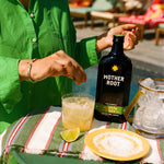 A person in a green shirt garnishes a jalapeño ginger drink with ice, lime, and salt. The scene is bright and summery, with a relaxed outdoor vibe.