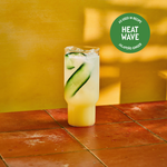 A tall glass of jalapeño ginger drink with ice and cucumber slices on a sunlit, terracotta-tiled surface. The warm yellow background evokes a summer vibe. Text reads “Heat Wave.”