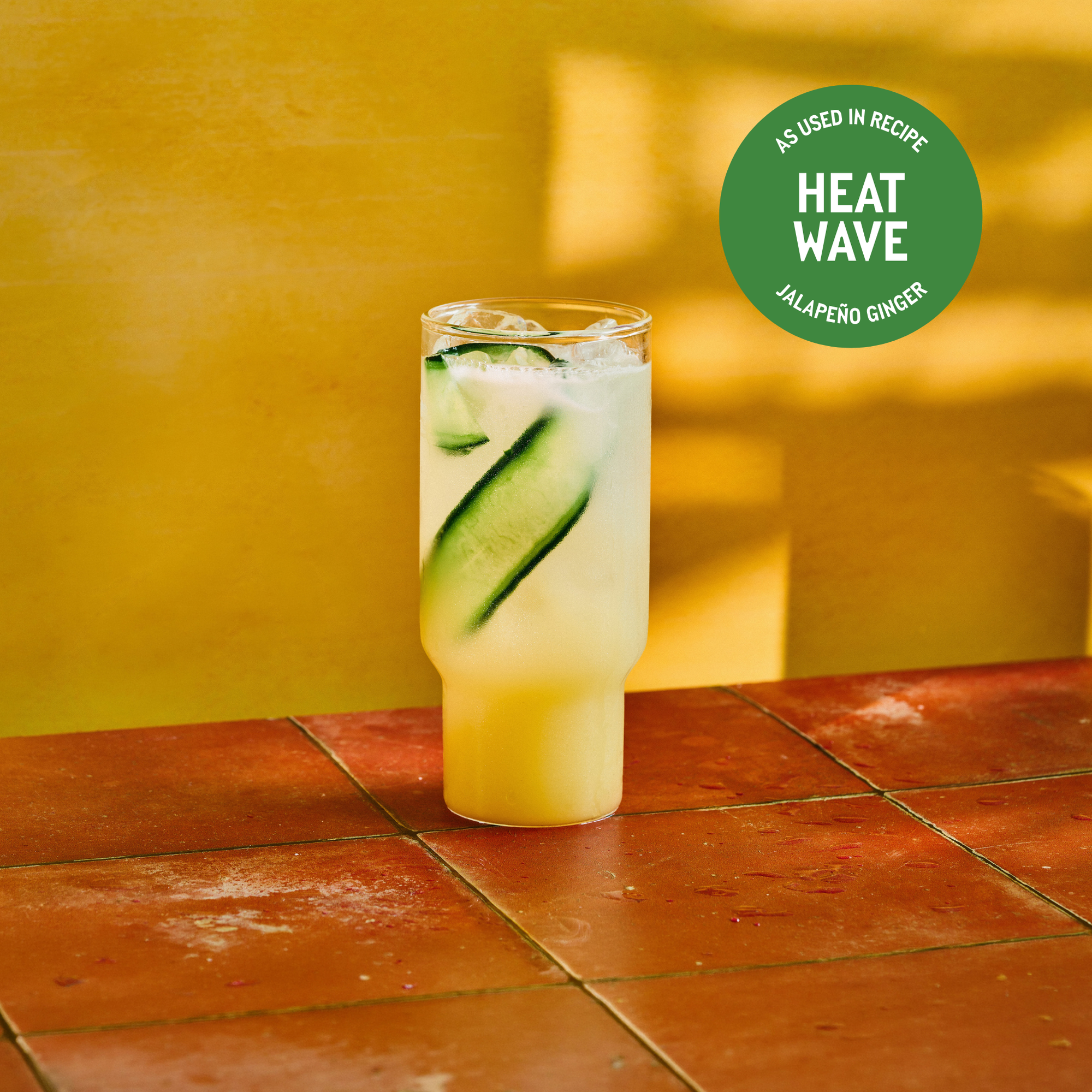 A tall glass of jalapeño ginger drink with ice and cucumber slices on a sunlit, terracotta-tiled surface. The warm yellow background evokes a summer vibe. Text reads “Heat Wave.”