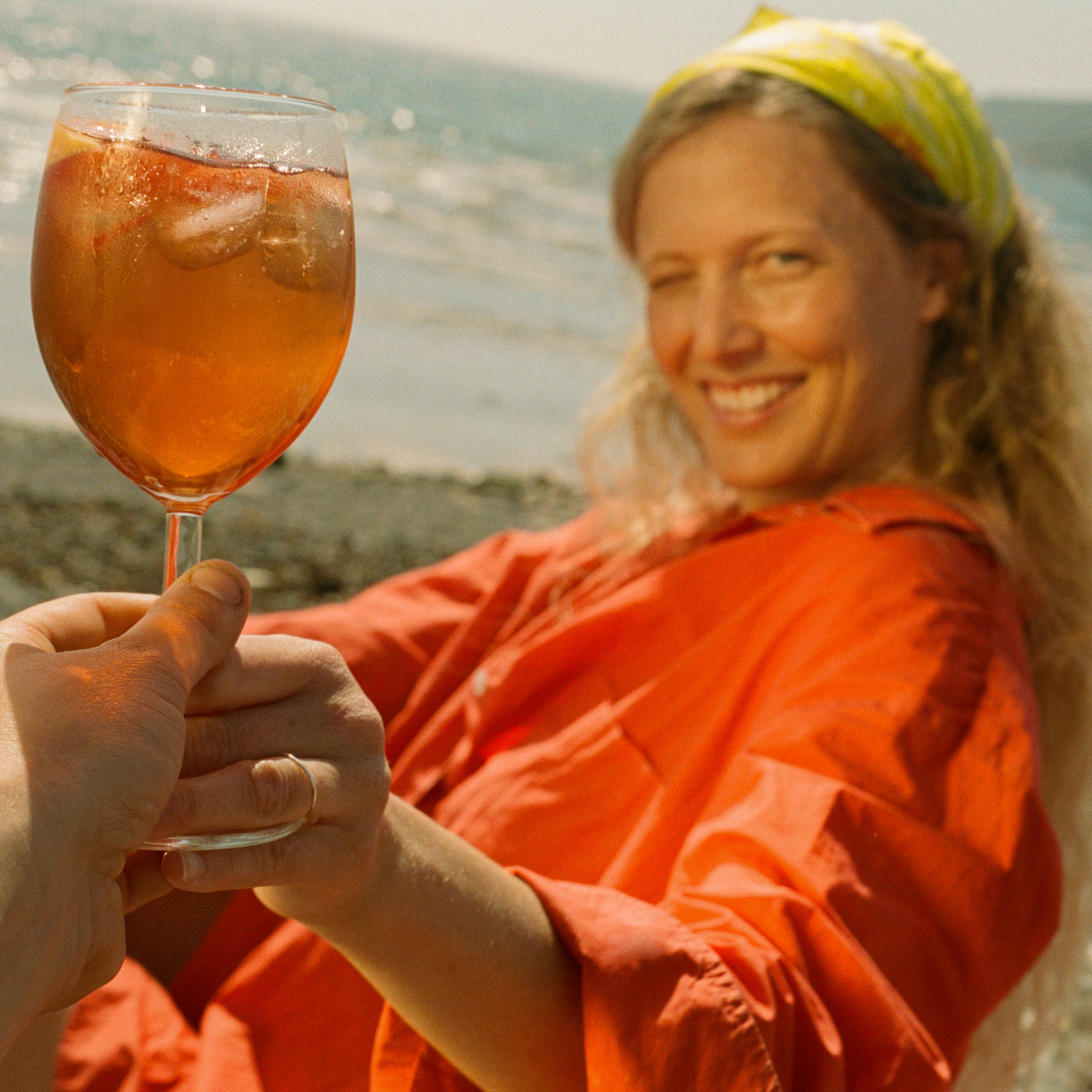 Smiling woman in an orange shirt and green headscarf holding up a Mother Root spritz over ice by the sea