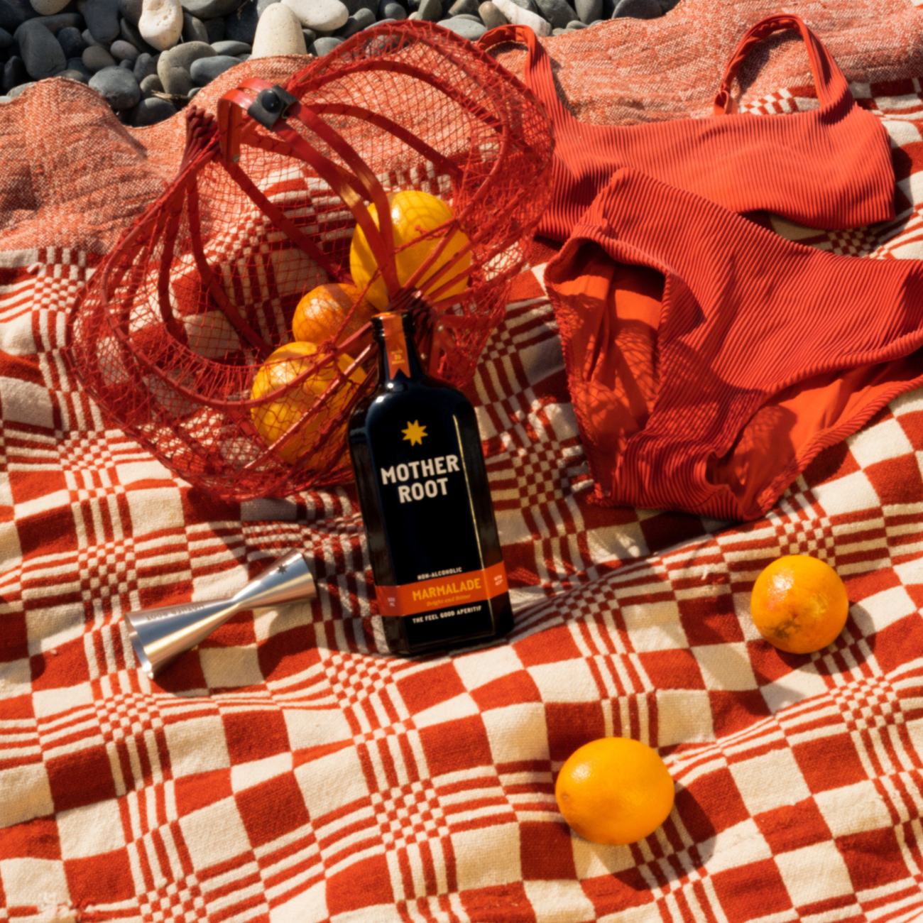Mother Root Marmalade non-alcoholic aperitif bottle on a red and cream checked picnic blanket, beside a red string bag of oranges, a stainless steel jigger and scattered citrus on a pebble beach